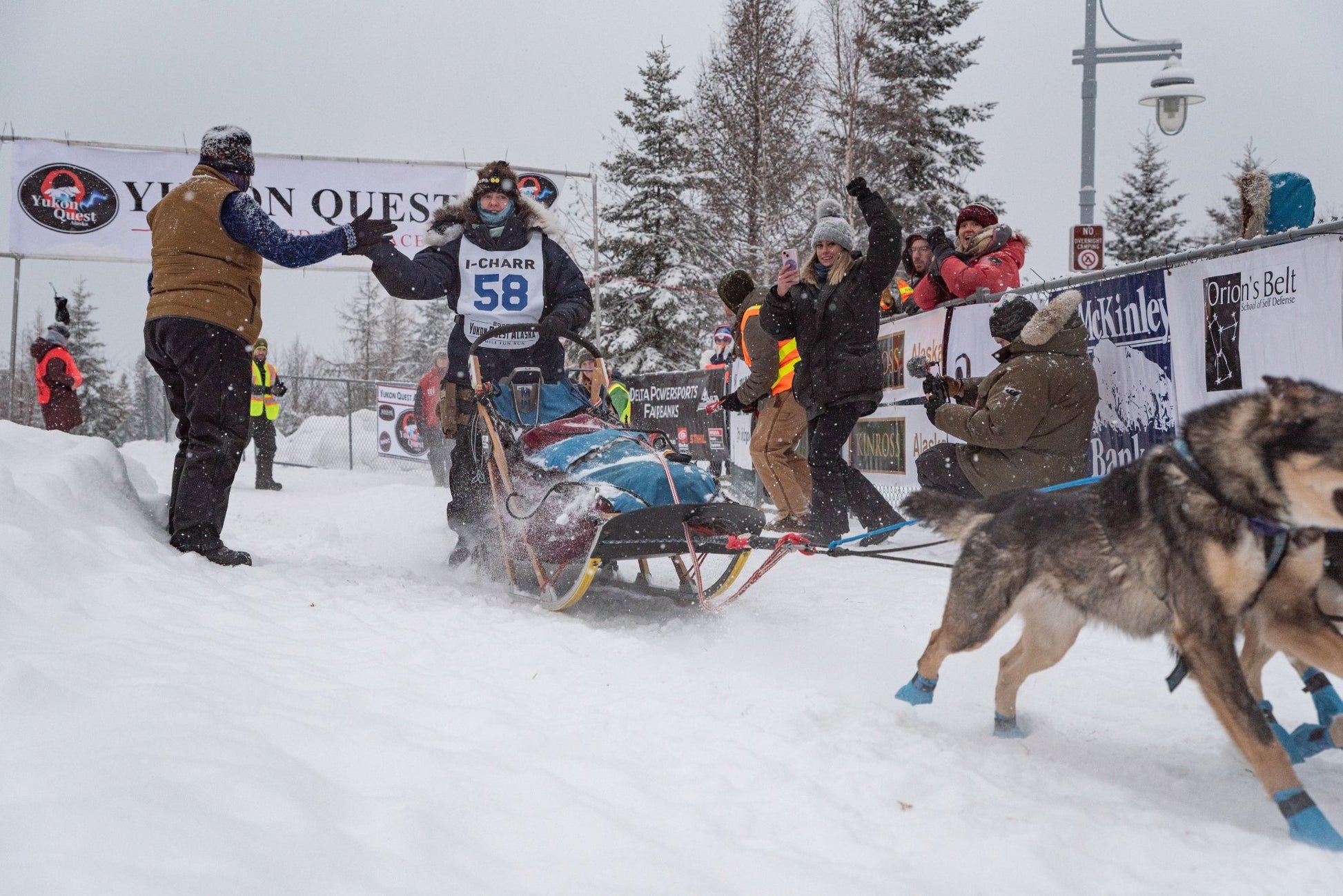 Dog sled race with musher and team in snowy landscape, spectators cheering.